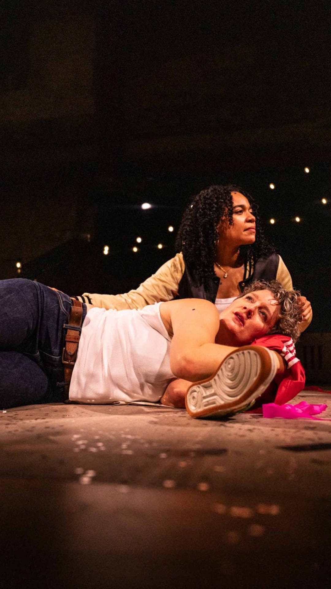 A person lies on the floor with their head propped up on a womans leg, the woman runs her hand through her hair and looks into the distance