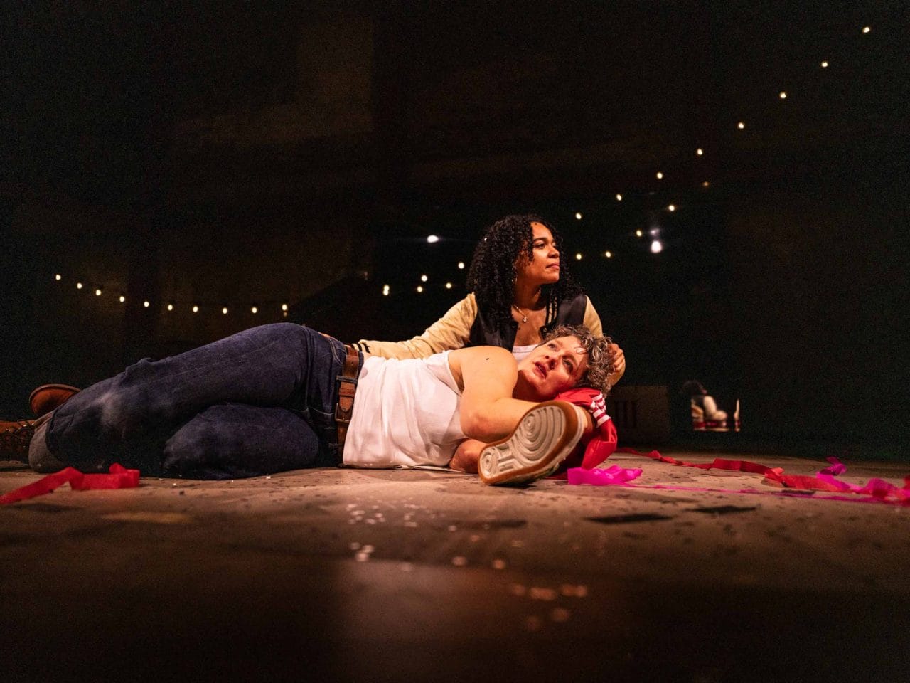 A person lies on the floor with their head propped up on a womans leg, the woman runs her hand through her hair and looks into the distance