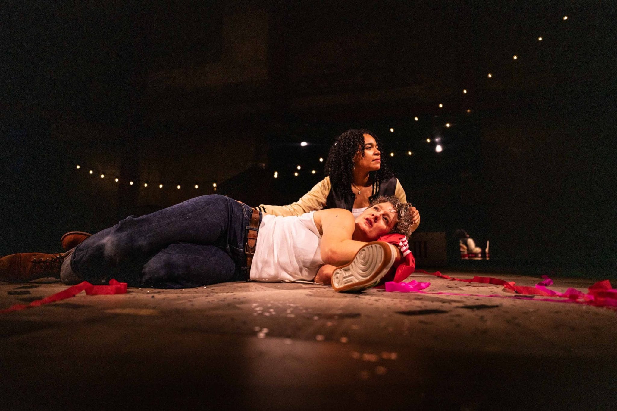 A person lies on the floor with their head propped up on a womans leg, the woman runs her hand through her hair and looks into the distance