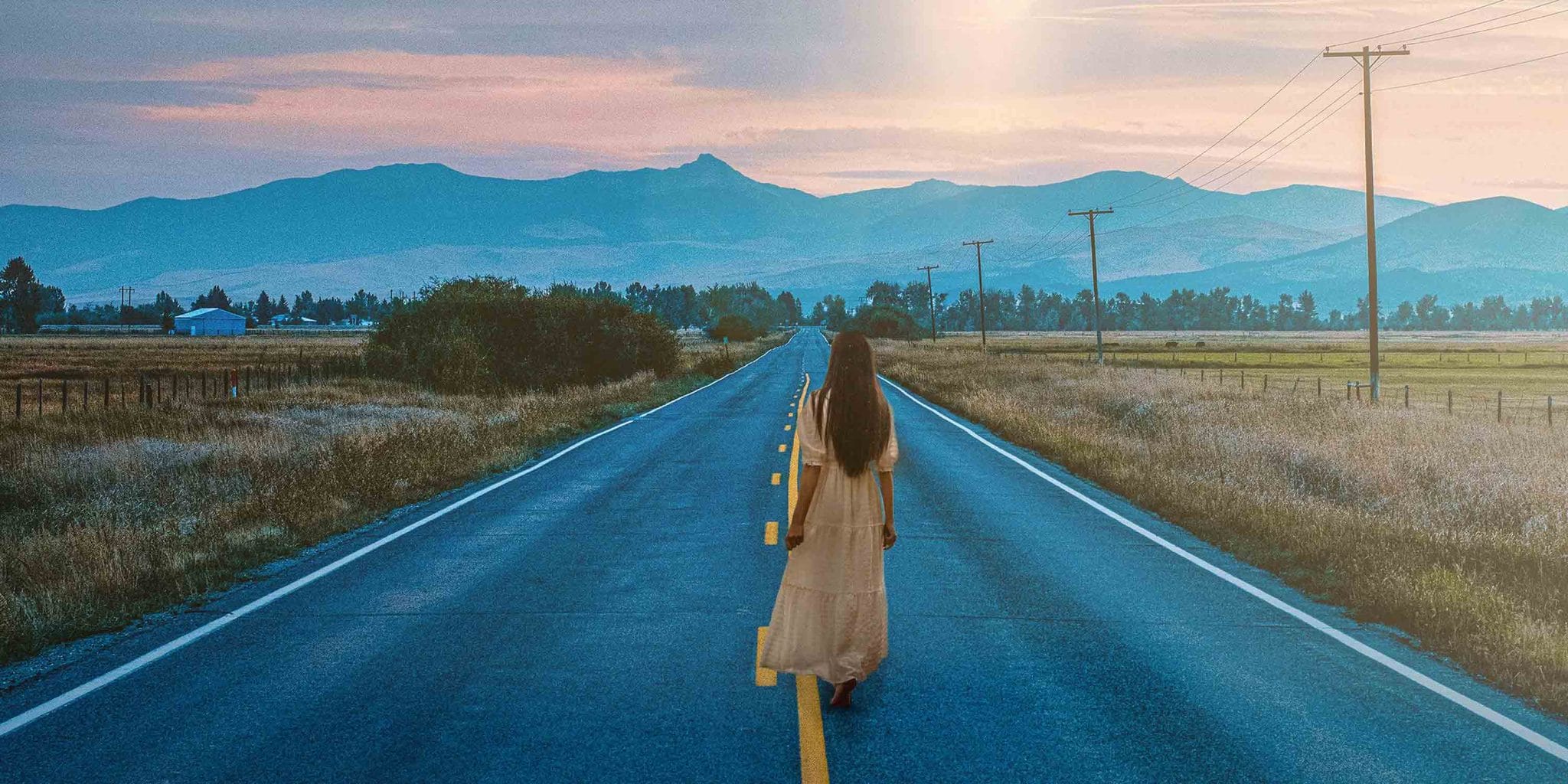 A woman is walking in a street with no shoes on that leads to a the hills.