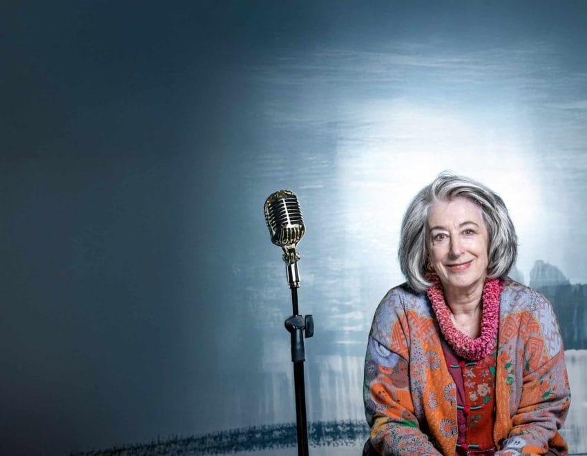 A photo of Maureen Lipman sitting wearing a colourful jacket. Next to her is a silver microphone.