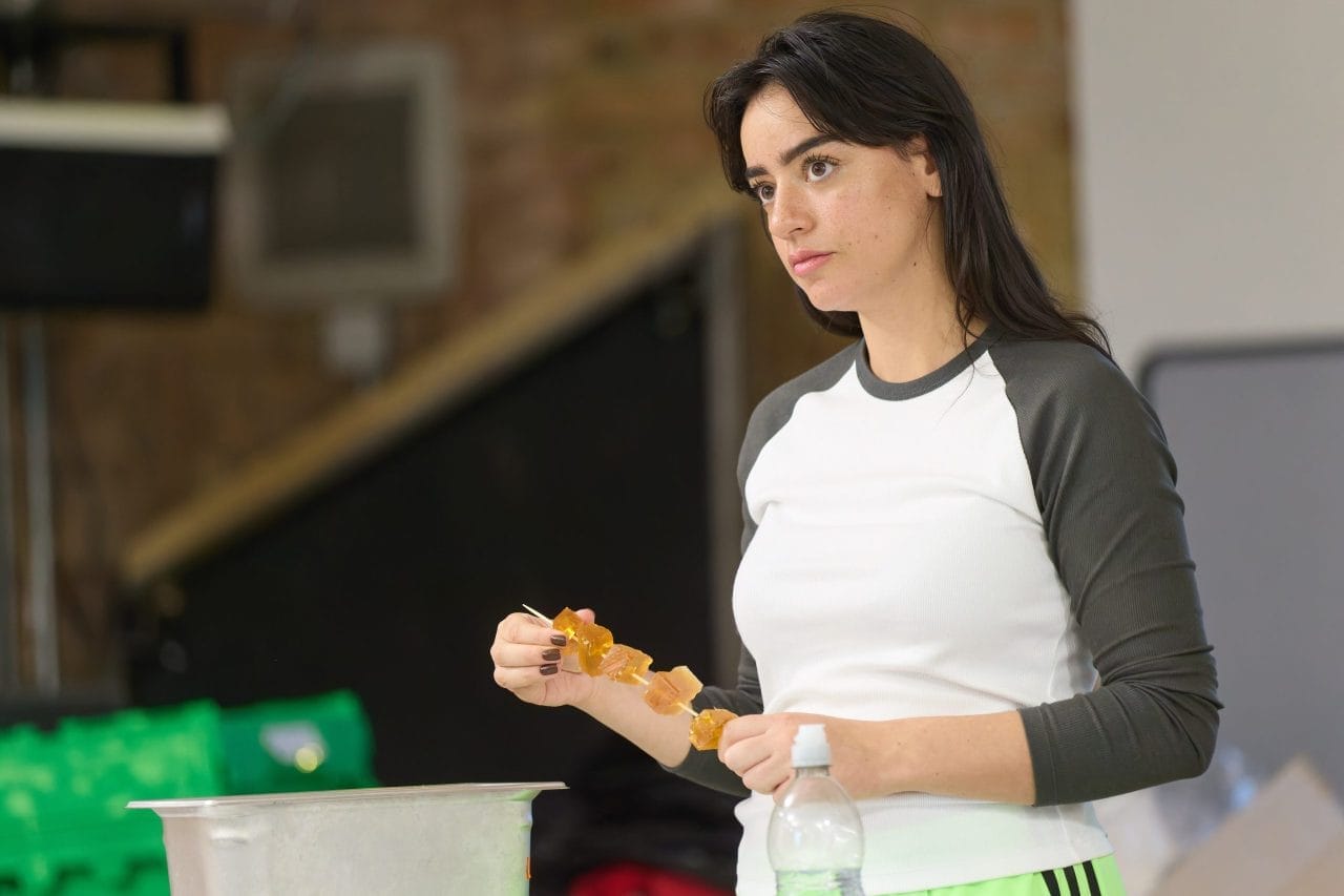 Mithra Malek wears a black and white baseball top as she puts fake chicken chunks onto a skewer. She looks into the distance with a concerned expression.