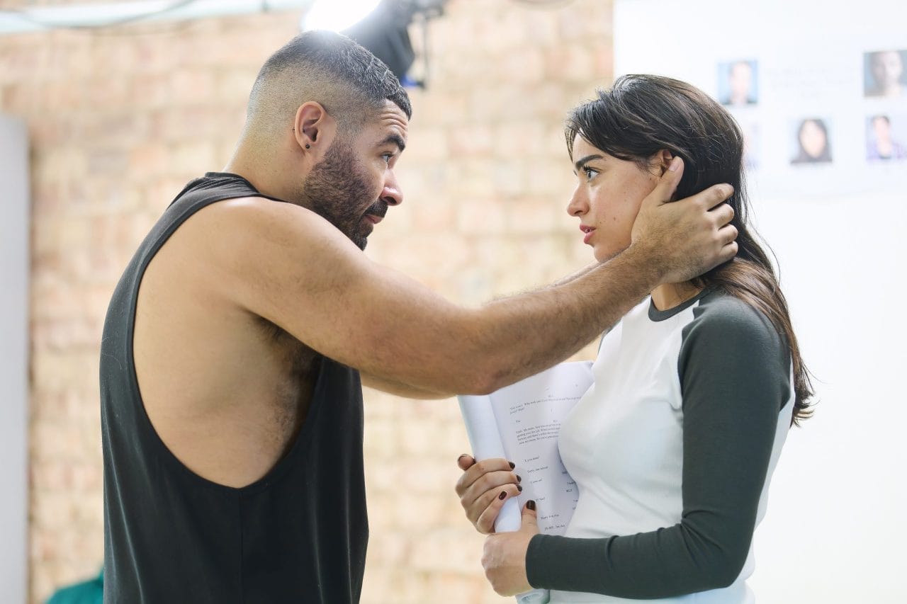 Ash Hunter is wearing a black tank top, reaching out to grip the side of Mithra Malek’s face and neck. She is wearing a black and white tank top and is holding her script. They both look concerned and serious.