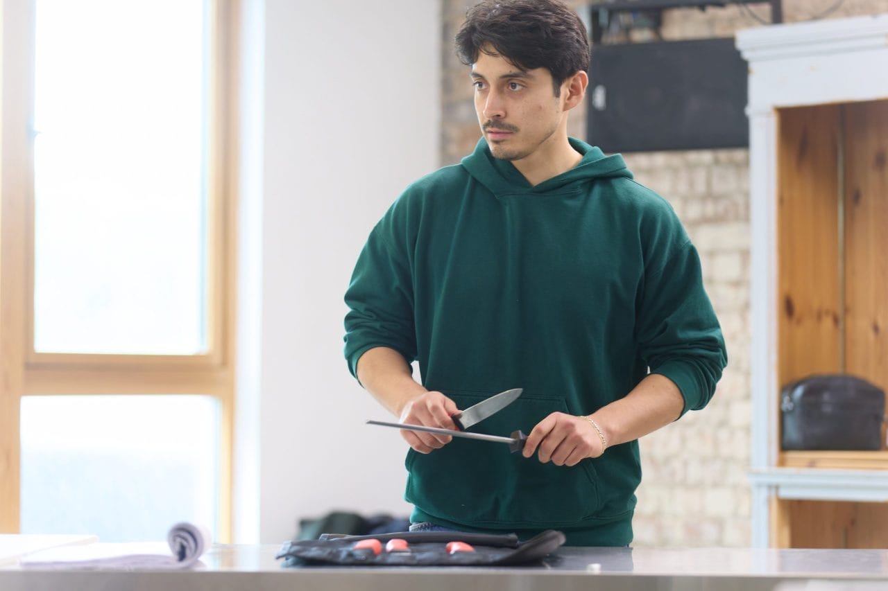 Marcello Cruz wears a turquoise hoodie with the sleeves rolled up. He has a serious expression while he sharpens a knife. The rest of the knives are in a knife roll on the table in front of him.