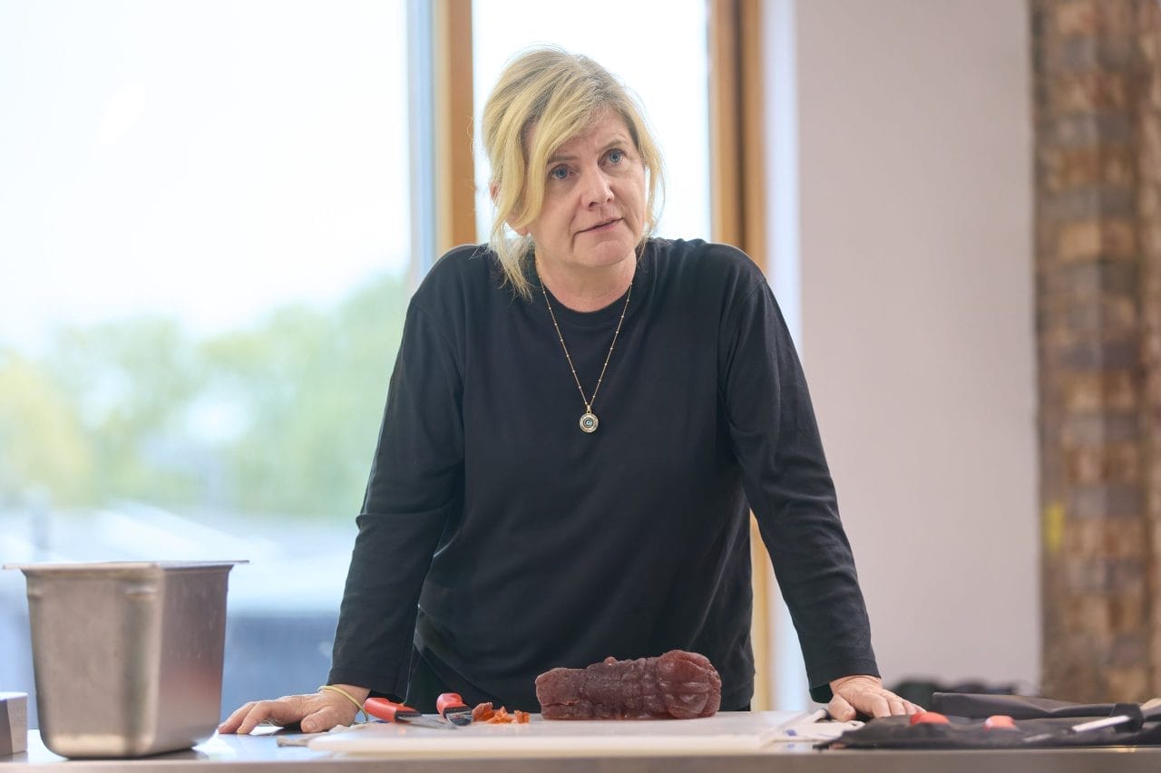 Jackie Clune wears a black long-sleeved top and black trousers and a gold necklace. She is standing at a silver metal table with a fake steak in front of her. On the table is a roll of knives and a metal bucket. Her hands are resting on the table, and she has a serious expression. 