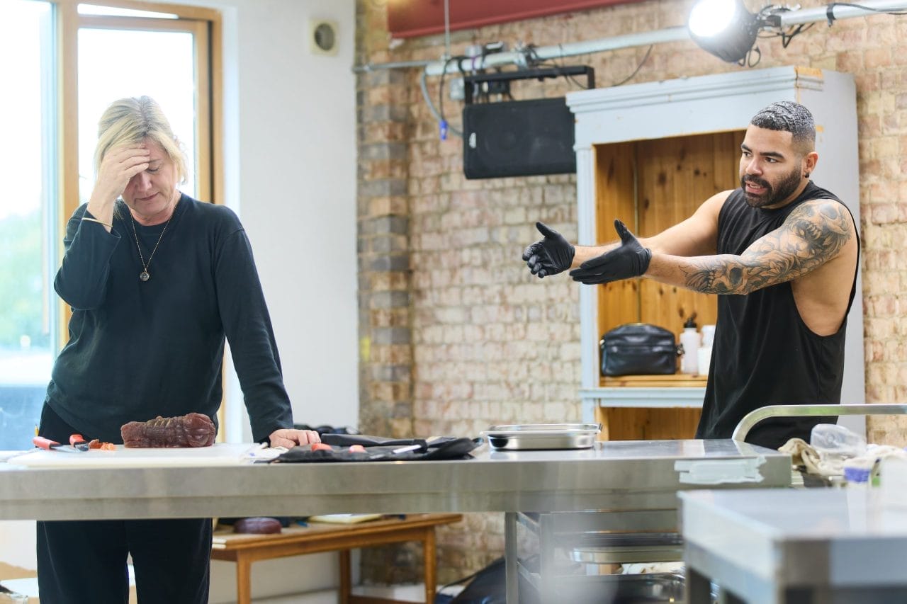 Jackie Clune wears a black long-sleeved top and black trousers and a gold necklace. She is standing at a silver metal table with a fake steak in front of her. She leans on the table with one hand, the other hand on her forehead and her eyes shut. Ash Hunter stands to her right behind the table wearing a black tank top with his tattoo sleeve visible, wearing black vinyl gloves. He has an angry expression and is gesturing to her with both hands.