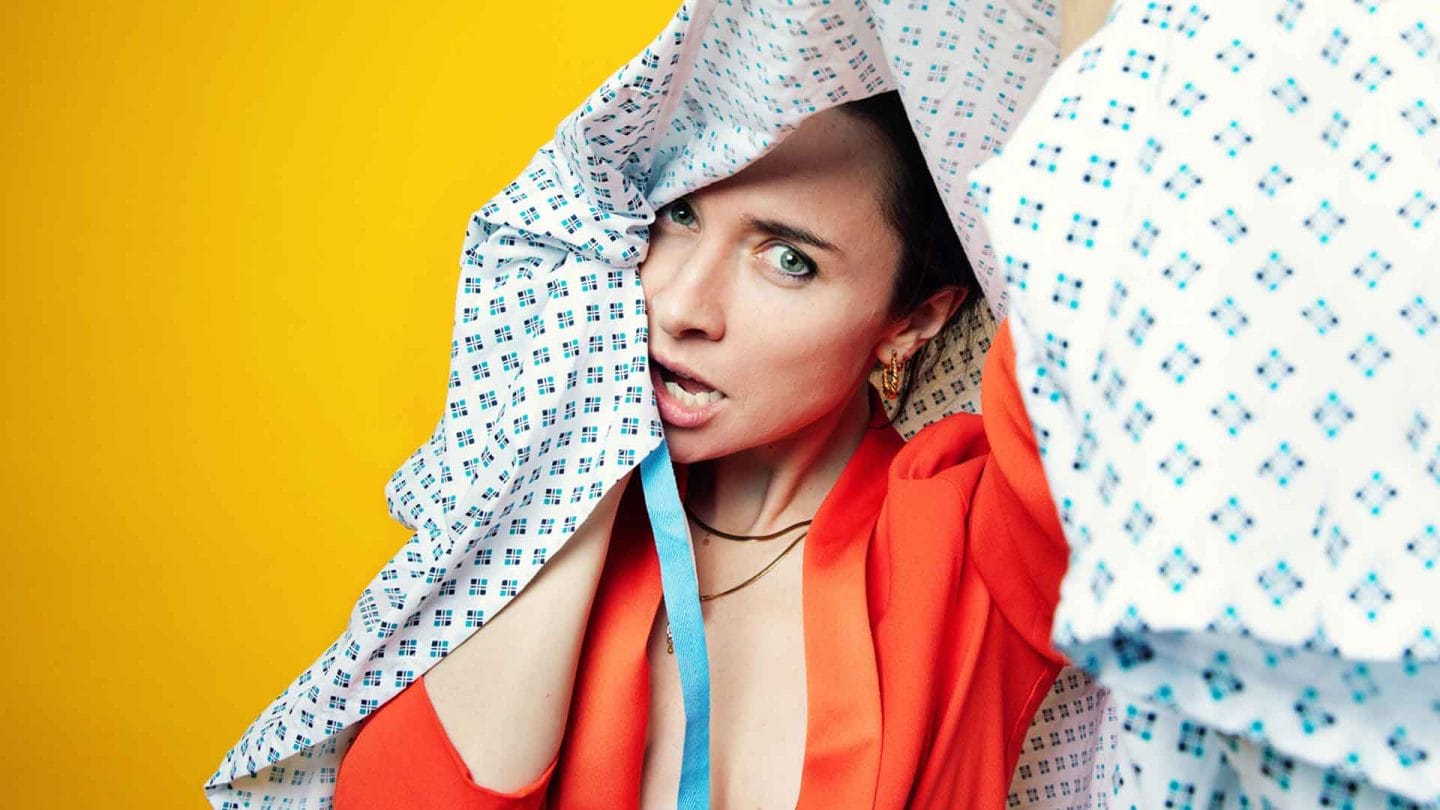 A woman wearing an orange blazer looks at us from under a hospital gown, as though she is tangled up in it. The background of the photo is yellow.