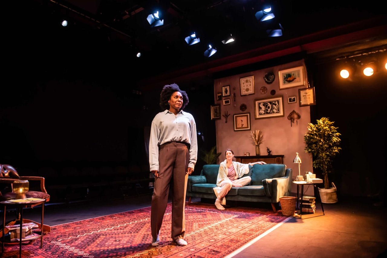 An image of a woman standing on stage on a rug and then another woman sitting on a sofa behind her looking at her