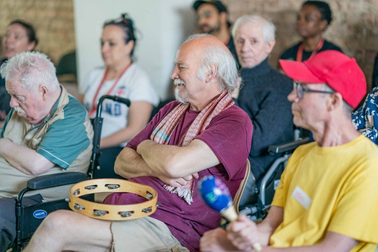 Photo of a man with a tambourine on his lap sitting next to other people with various instruments.