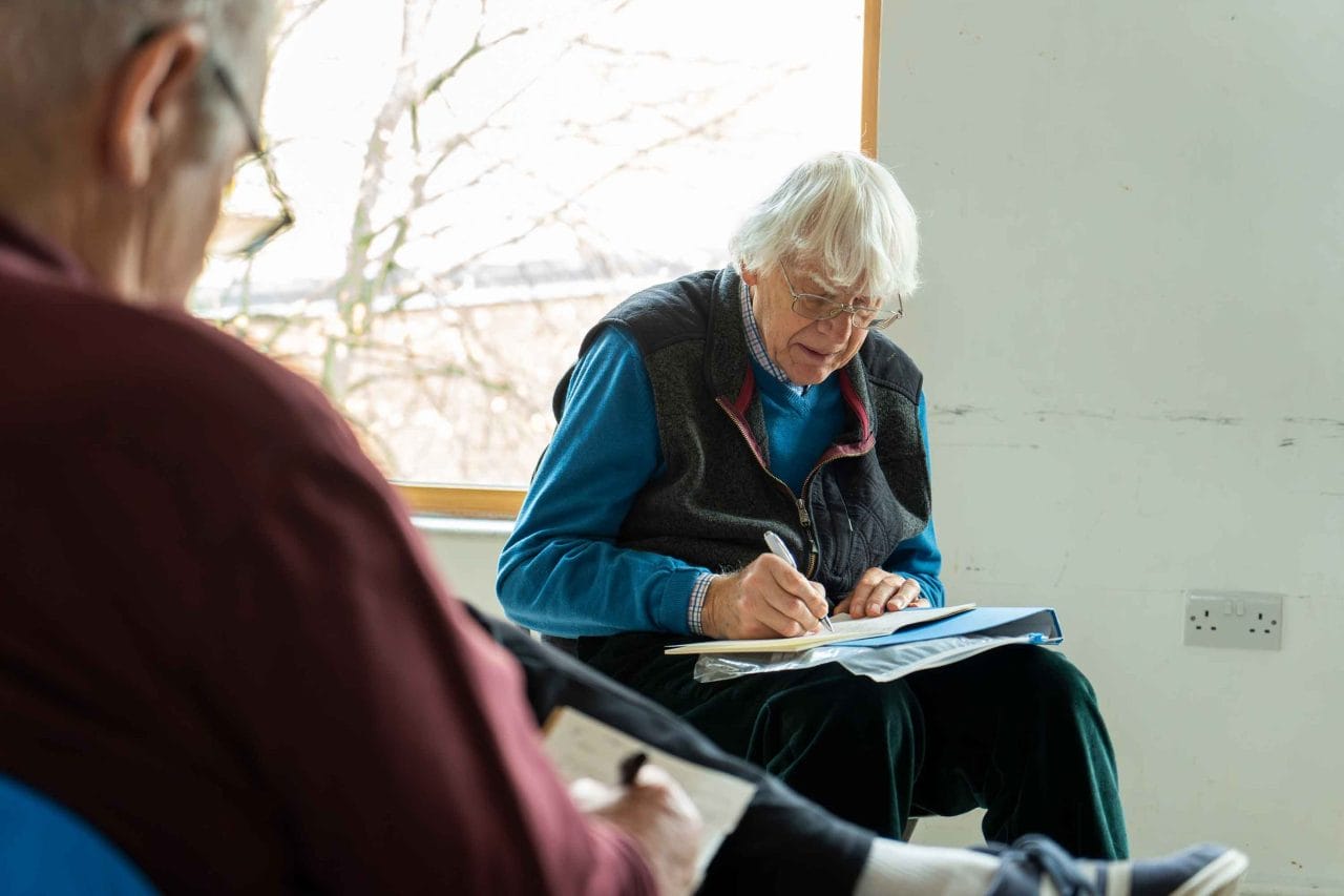 A man in a sweater and glasses writes in a notebook with a pen while sitting in a chair.