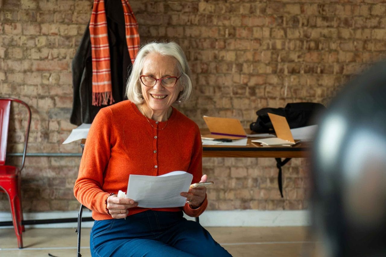 A woman sitting on a chair wearing a red sweater and glasses, holding a piece of paper, and smiling.