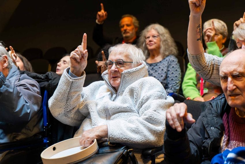 Photo of a person wearing a warm coat, playing with a tambourine with one finger up in the air.
