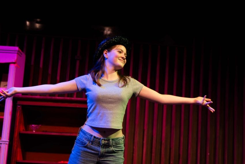 Photo of a girl on stage wearing a black hat with her arms out addressing the audience.