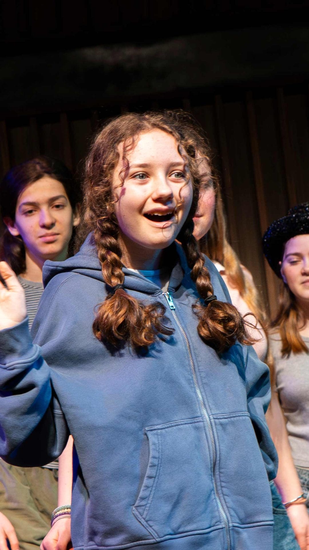 Photo of a young girl in a blue sweatshirt and two braids smiling and waving while on stage