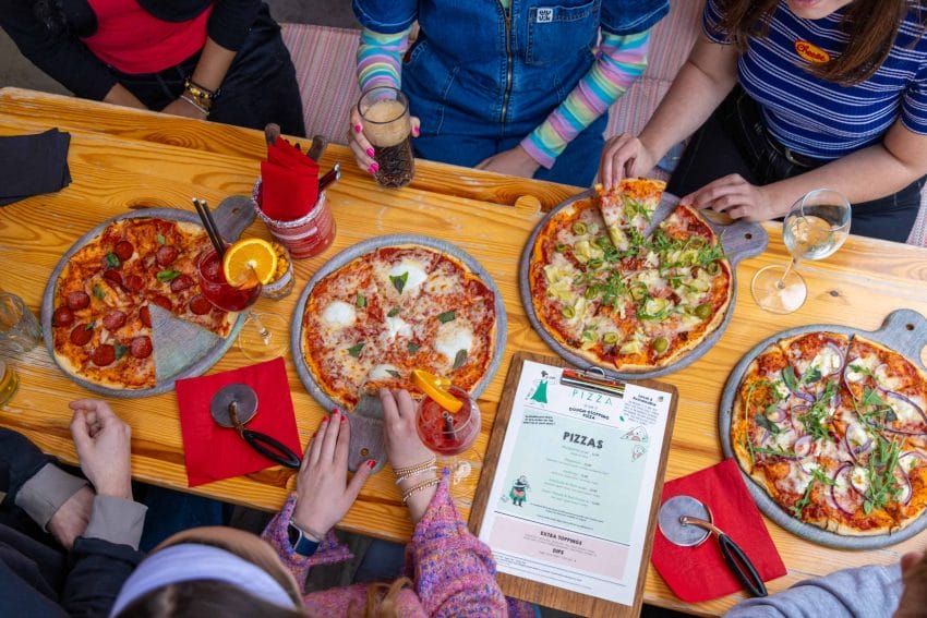 An image of a table with 4 Pizza's, drinks and 6 people sitting around the table