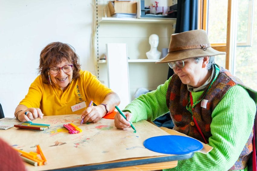 2 people sitting at a table smiling and coloring with green and purple markers.