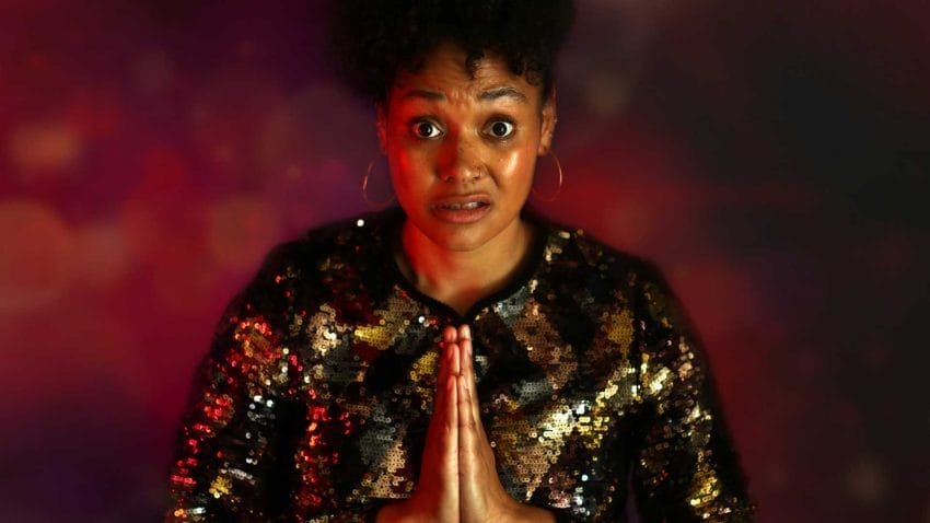 A woman in a sparkly shirt looks at the camera with a bewildered expression on her face. She holds her hands to her chest in prayer.