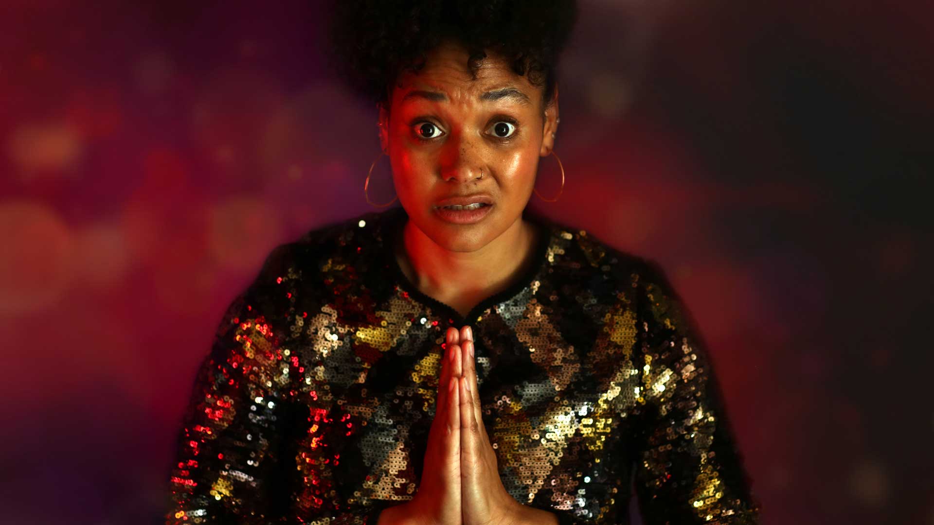 A woman in a sparkly shirt looks at the camera with a bewildered expression on her face. She holds her hands to her chest in prayer.