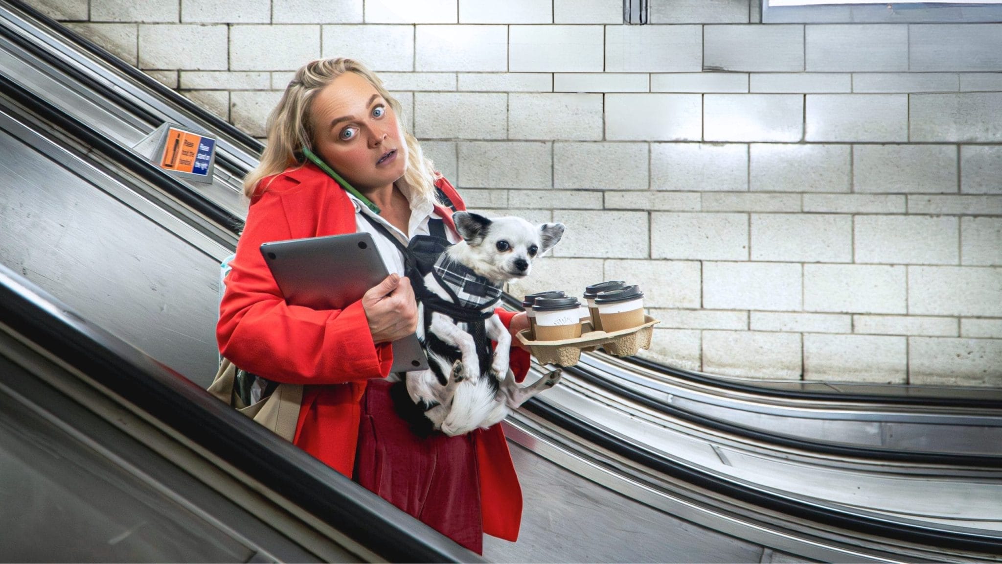 A woman holds a dog, a laptop and coffee, and struggles with holding a phone to her ear. She wears a red blazer and stands on an escalator.