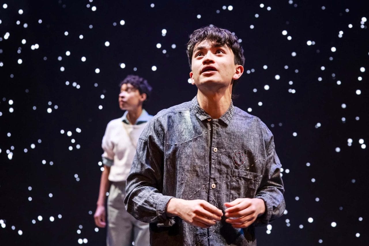 A man stands looking up. In the background behind him, someone else looks ahead of them. The backdrop shows stars on a black screen.