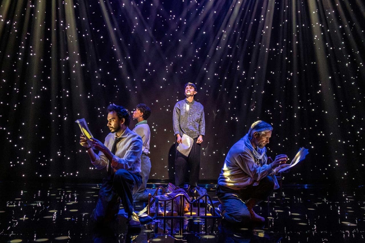 Four people stand holding pieces of paper. The back wall is covered in stars and strobes of light.