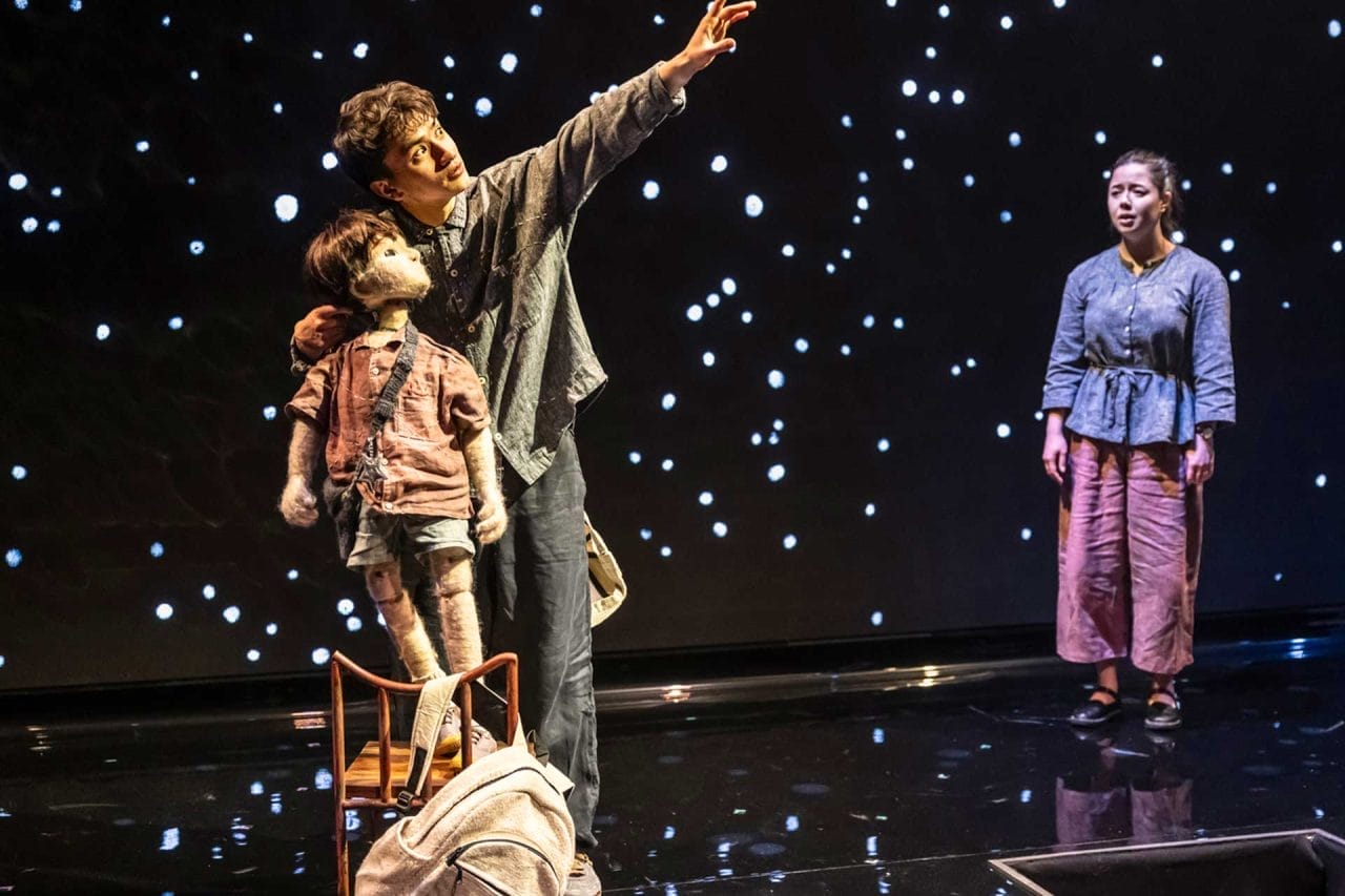 A man hold a puppet on a chair and points to the sky. In the background are stars.