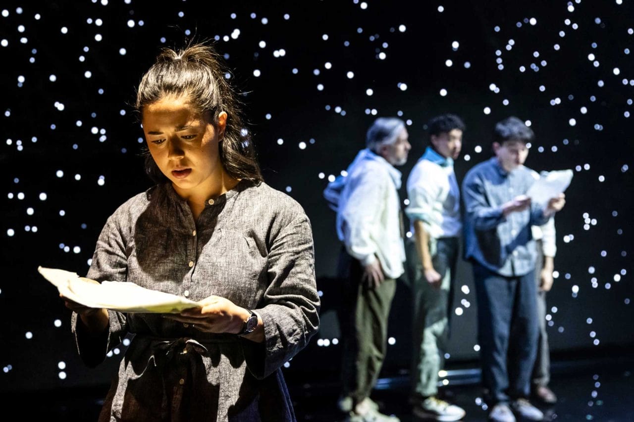 A woman holds a piece of paper in front of her. In the background, three people crowd round a piece of paper. The back screen is covered in stars.