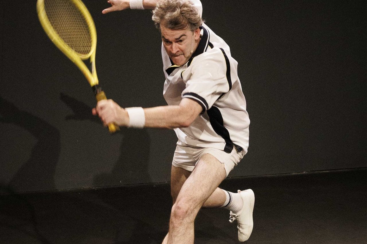A still of a man, in his 40s, in a white tennis outfit, with a yellow racket, ´going for a shot´ leaning forward on one leg in the process.