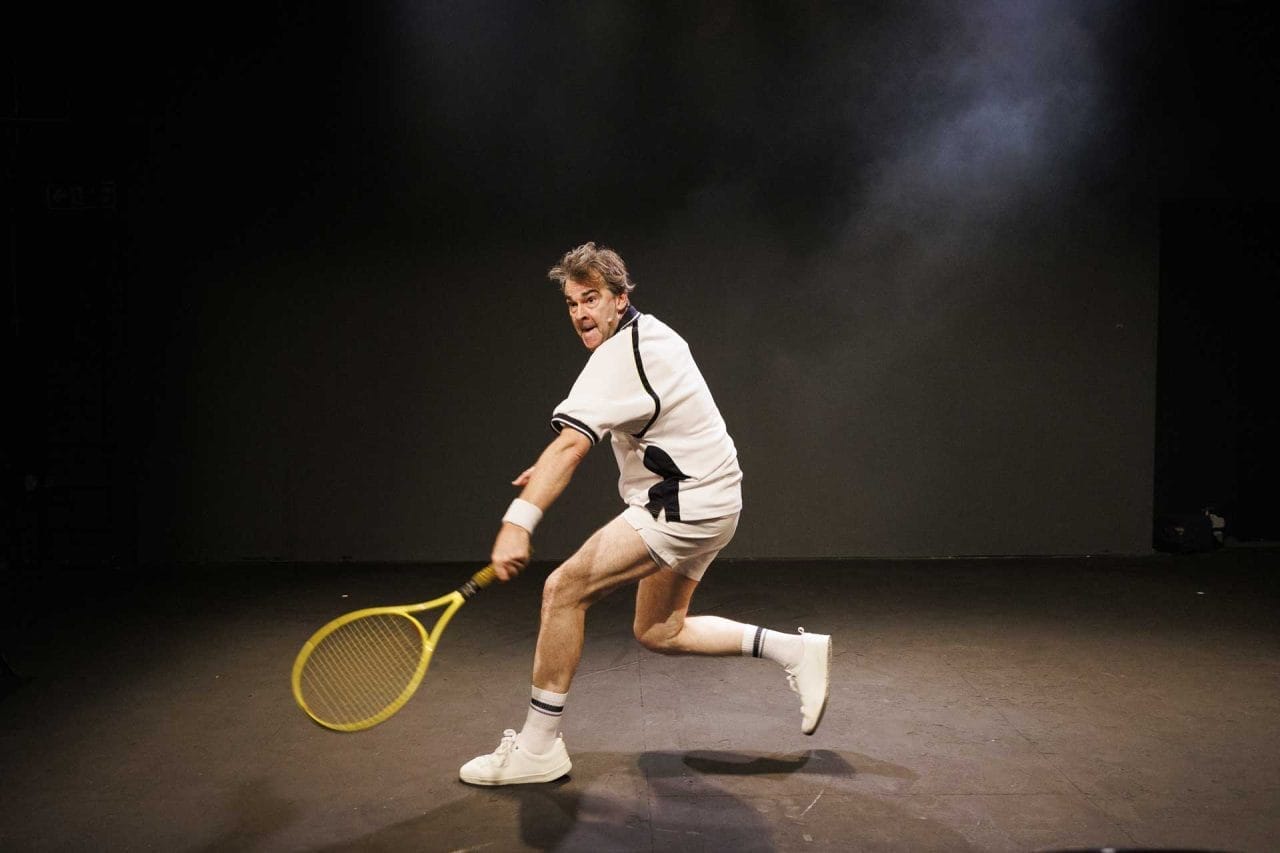 A still of a man, 40s, in a white and navy tennis shirt and white tennis short, socks and shoes, with a yellow tennis racket in his left hand, ´going for a shot´. He has a spotlight on him, and with a black background.