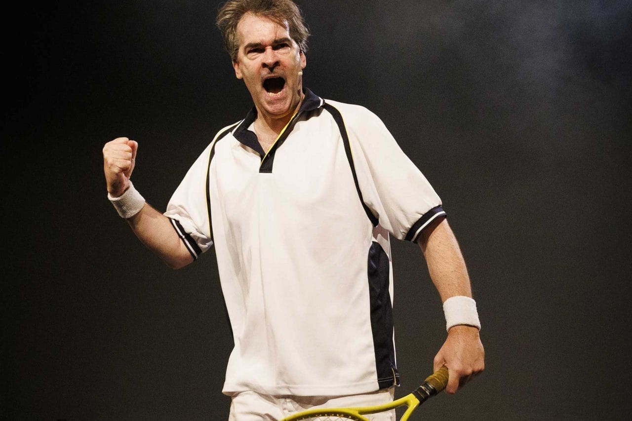 A man, in is 40s, in a white and navy tennis shirt and white shorts, fills the photo, celebrating winning a tennis ´point´, as holds a yellow tennis racket in his left hand.
