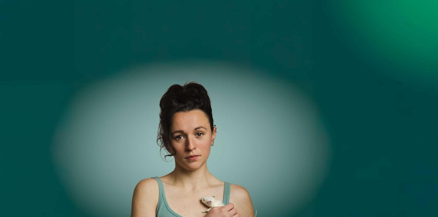 A woman in a green tank top holds a mouse to her chest. She looks out with a serious expression on her face.
