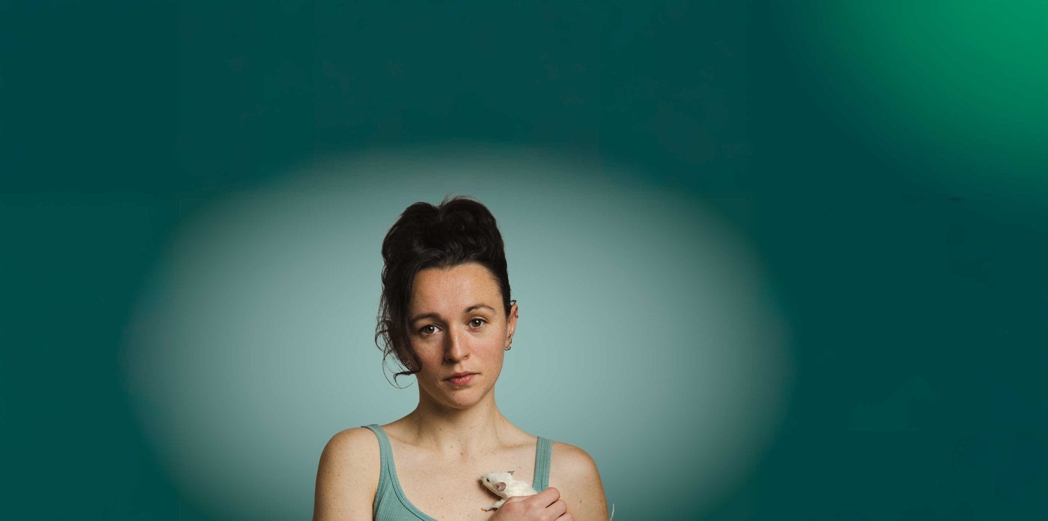 A woman in a green tank top holds a mouse to her chest. She looks out with a serious expression on her face.