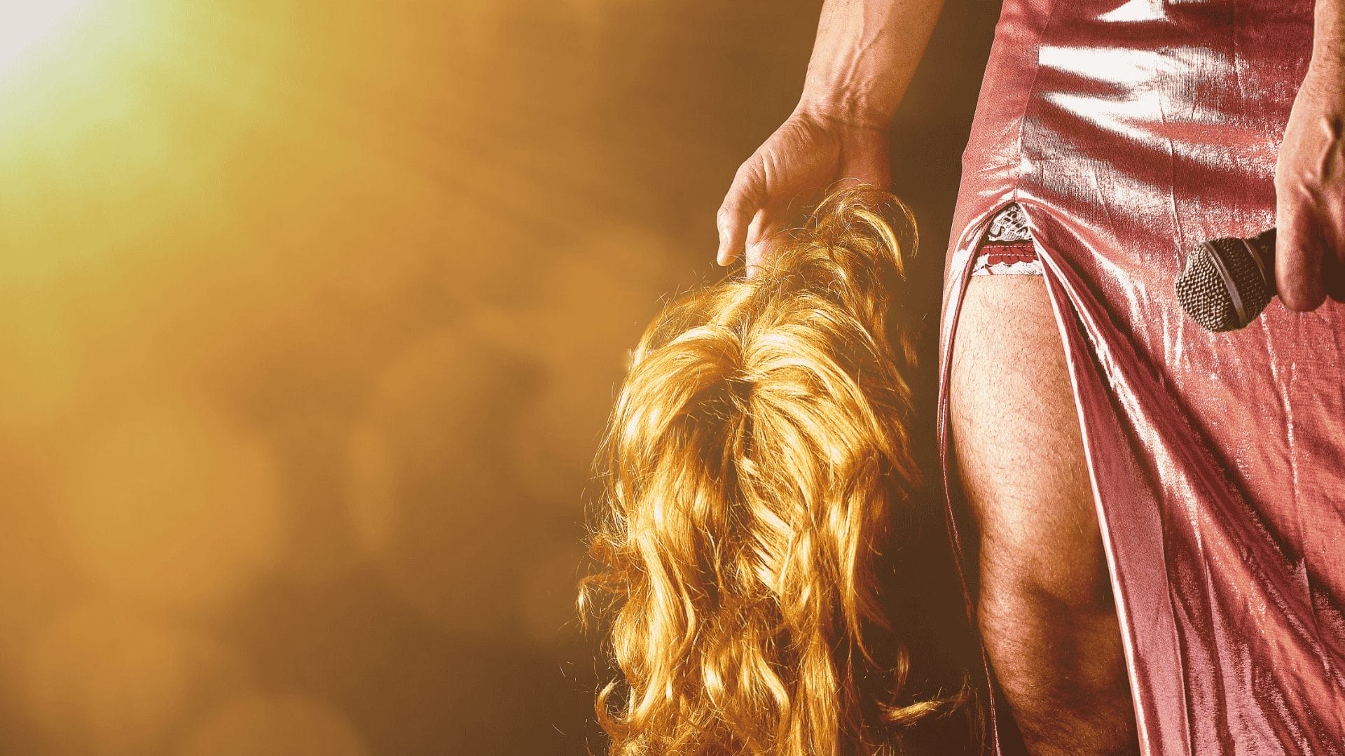 A bright orange light is seen coming from the top left hand corner, shining onto a long blond wig, held by a more masculine hand. The right third shows someone in a pink dress holding a microphone. A split in the dress reveals the person's thigh - which is also more masculine.