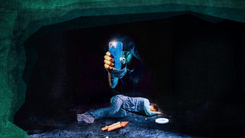 Hooded person is unclearly seen, as he holds his phone with the torch is on, in his right hand, as there is a body lying face turned down below him. Surrounded by what looks like a green cave.