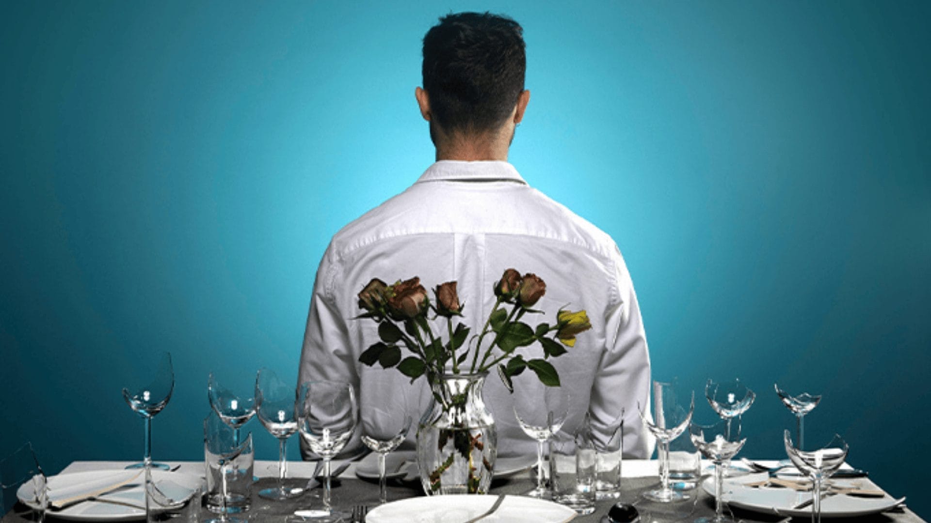 Man wearing dress shirt facing back-facing. Behind him is a table, where there is a vase of dead flowers, along with 4 plates, cutlery, and some wine glasses, as well as regular water glasses. A few of the wine glasses on one side of the frame have the bowls smashed.