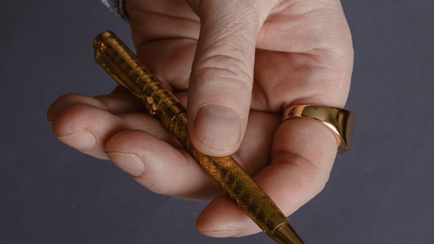 Close up focus on a masculine left hand, with a gold signet ring on the index finger. The hand is holding a golden pen. The finger names are very short.