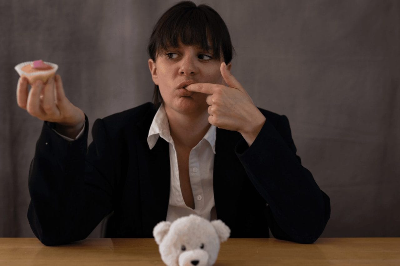 Woman in a central position, sitting down at a table where a small white teddy bear sits in front of her, as she looks to the right. She is licking her right index finger, and has a cupcake with icing.