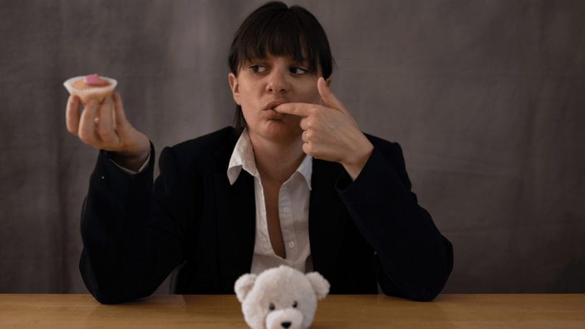 Woman in a central position, sitting down at a table where a small white teddy bear sits in front of her, as she looks to the right. She is licking her right index finger, and has a cupcake with icing.
