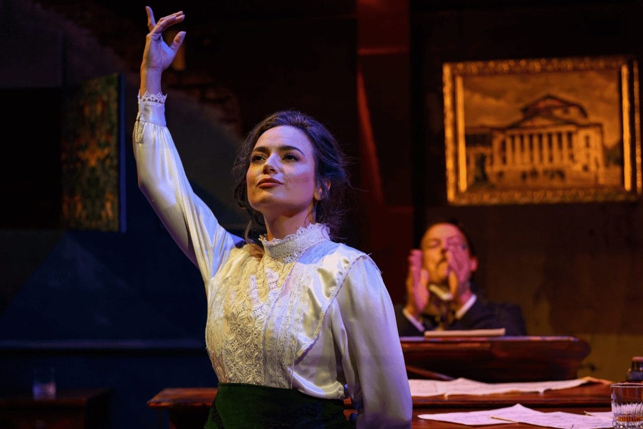 A woman with dark hair, a smart white shirt and a navy skirt has her right hand up in the air, and left hand behind her back, holding herself as if she has just done something very successfully. Behind her, there are 2 tables with scattered pieces of paper on them, a small glass with some sort of alcohol in, and a the blur of a man in the background, clapping. A picture can also bee seen hanging up behind the man - of a big old building, such as an opera house, in a golden frame.