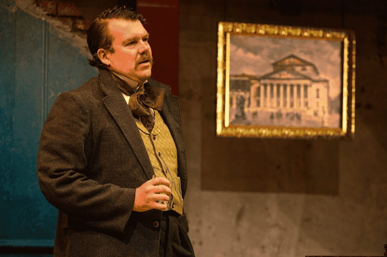 A middle aged looking man with dark brown hair and mustache, matching his jacket and trousers, and cream waistcoat looking across the stage, clutching a glass with his right hand. In the background is a framed picture of a big building with pillars, such as an opera house. The frame is gold colored.