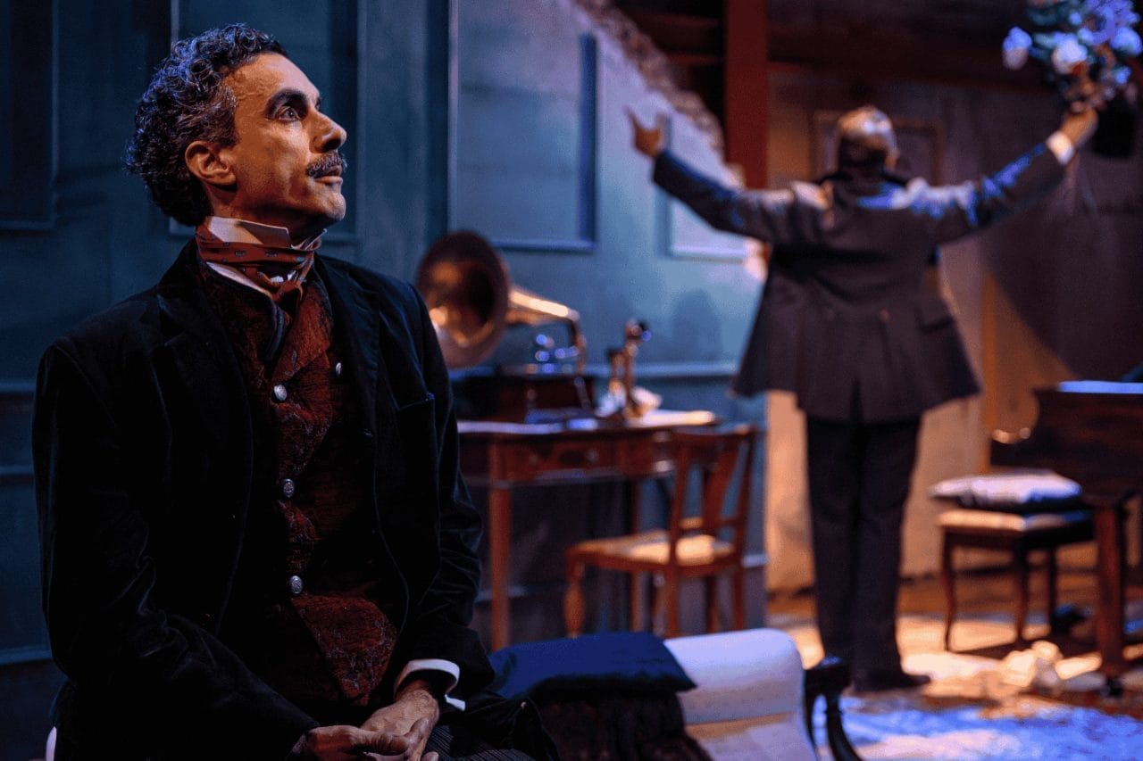 A man with dark grey hair and a mustache, in a black jacket and grey trousers is sitting on a white chaise lounge, looking out into the 'world' with a serious tone, as if he was thinking a serious thought. Slightly blurred in the back, there is a brown chair and table, with a phonograph sitting on top and then also the end of a piano next to a piano chair and cushion. In between the piano and phonograph, a man in a dark suit faces away, with his hands spread out and up, with a bunch of flowers in his right hand.