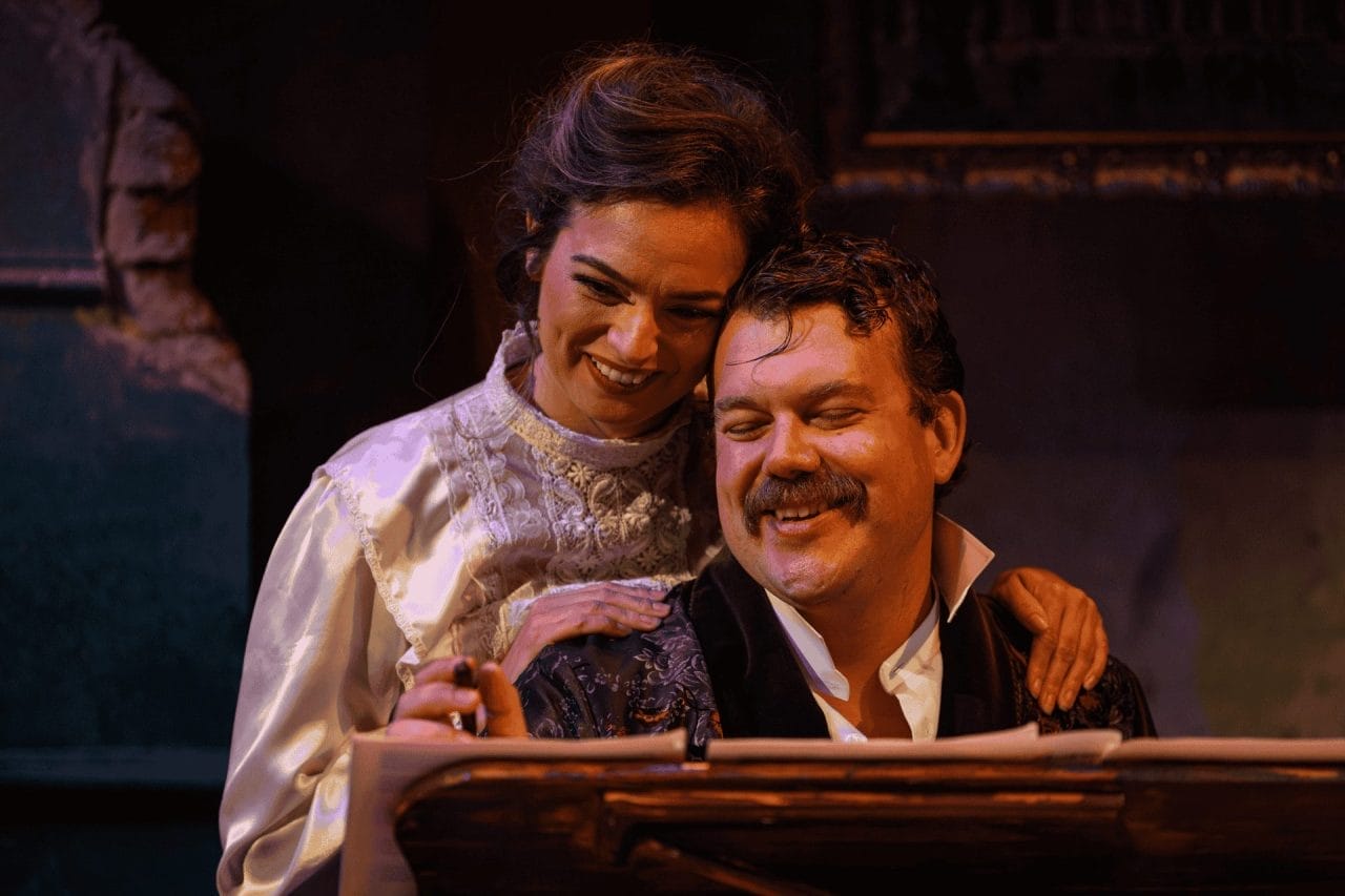 A man sits at a piano, looking exhausted, yet happy, as he has a pen in his right hand, as if to adjust some notes on his music sheet. He has an open collar and a black mustache. A woman, also smiling, is sitting behind him as she looks at his hand with the pen.