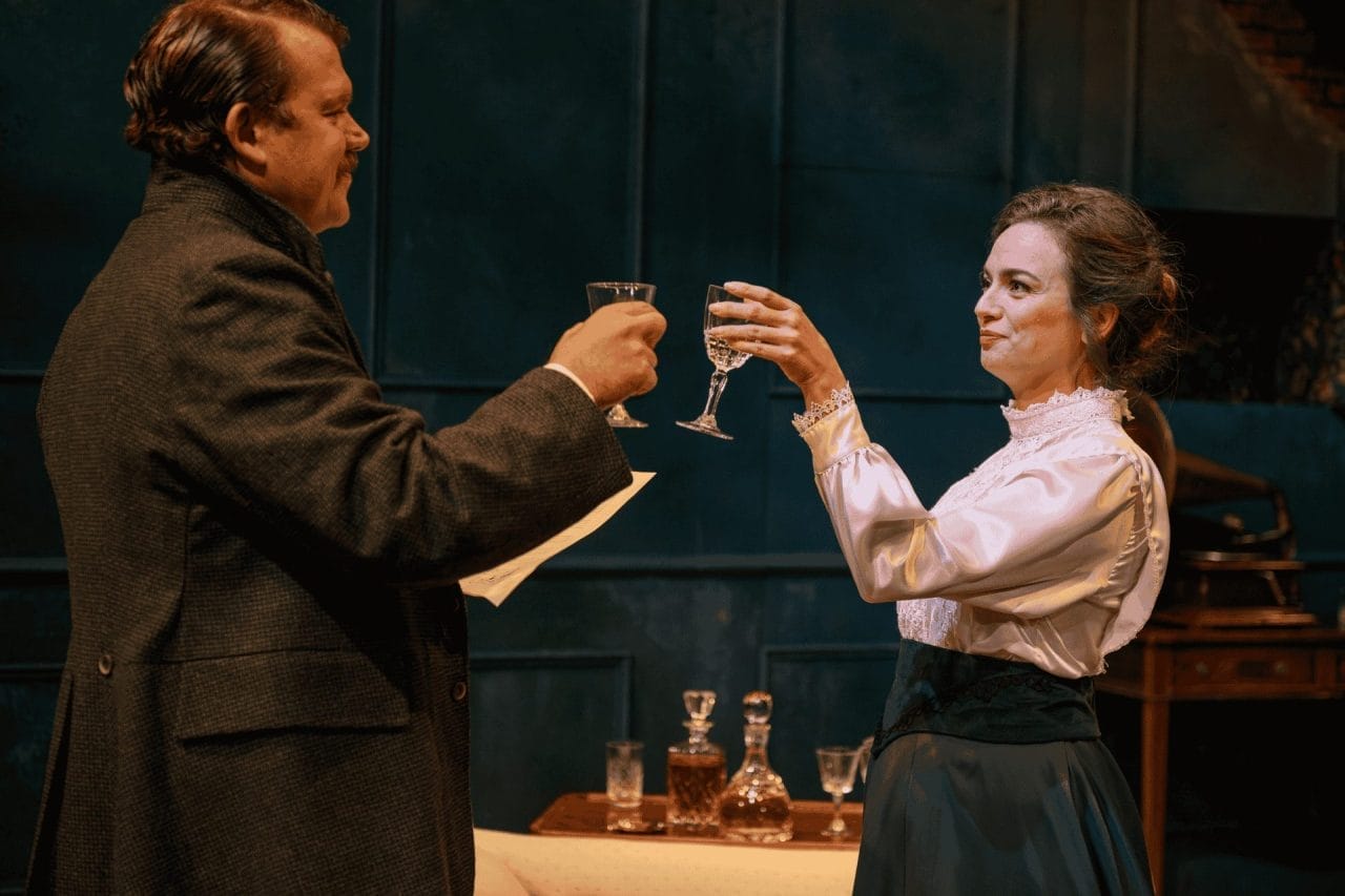 A man and woman are facing each other with a little smile, as they are in the process of clicking their glasses together. The man, who is tallker, is wearing a dark brown suit jacket with his collar up, with a piece of paper in his left hand (that is unreadable). The woman, wearing a very nice shirt and navy skirt, has a glass in her left hand. In between the two of them a small drinks table can be seen with two 19th century bottles seen of some sort of spirit.