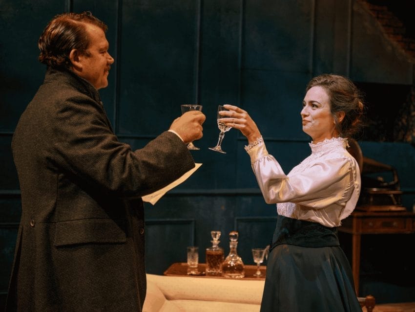 A man and woman are facing each other with a little smile, as they are in the process of clicking their glasses together. The man, who is tallker, is wearing a dark brown suit jacket with his collar up, with a piece of paper in his left hand (that is unreadable). The woman, wearing a very nice shirt and navy skirt, has a glass in her left hand. In between the two of them a small drinks table can be seen with two 19th century bottles seen of some sort of spirit.