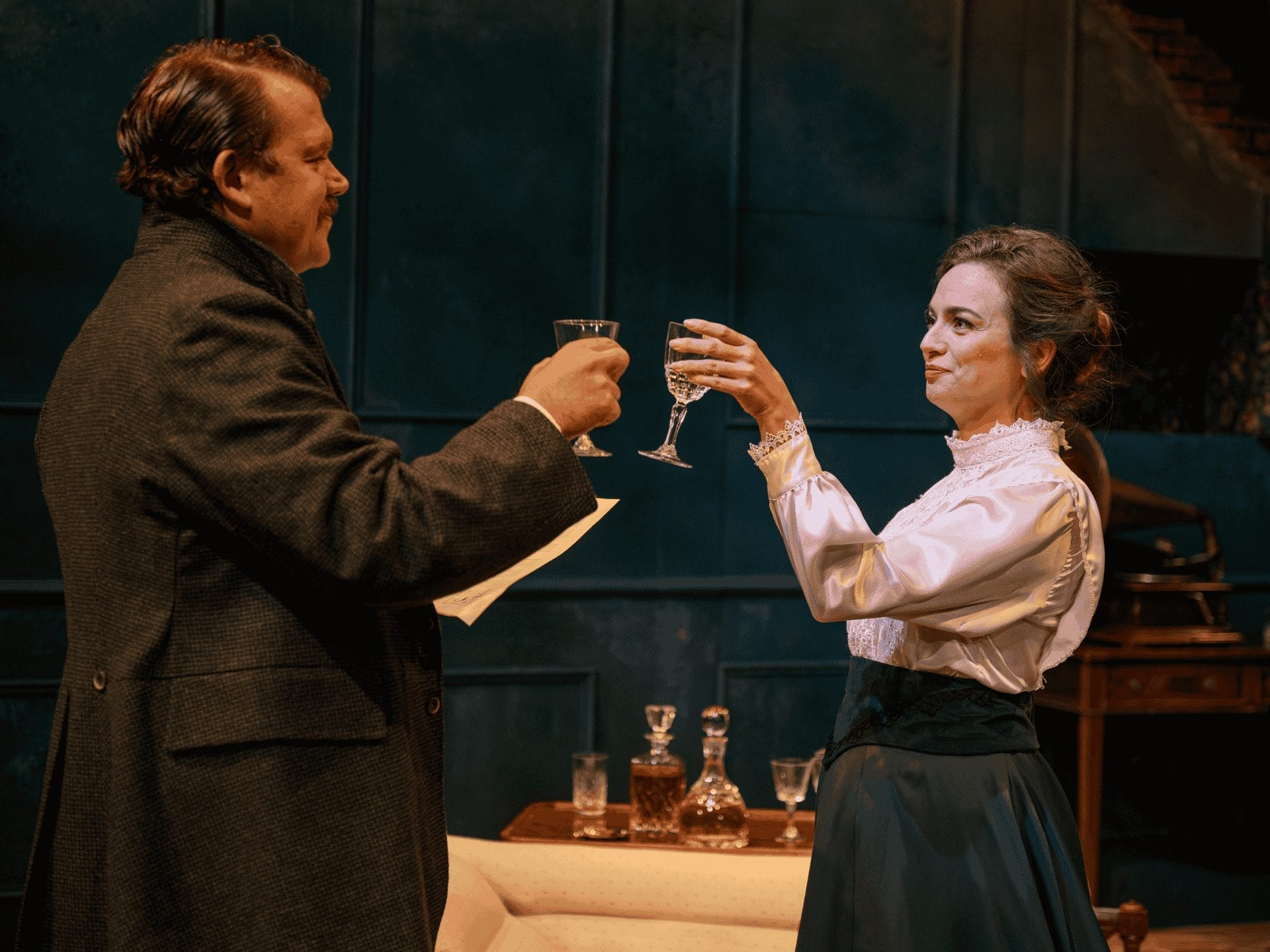 A man and woman are facing each other with a little smile, as they are in the process of clicking their glasses together. The man, who is tallker, is wearing a dark brown suit jacket with his collar up, with a piece of paper in his left hand (that is unreadable). The woman, wearing a very nice shirt and navy skirt, has a glass in her left hand. In between the two of them a small drinks table can be seen with two 19th century bottles seen of some sort of spirit.