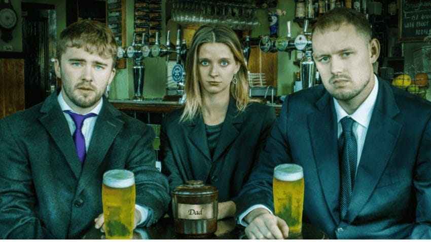 3 people sit in a pub, 2 men on either side, with a woman in the middle, all looking serious. The 2 men have a pint of beer in front of them, whilst the woman has a brown urn, reading 'Dad' on the front. Behind the trio, the actual bar can be seen, with a number of taps, glasses and other bottles on a shelf.