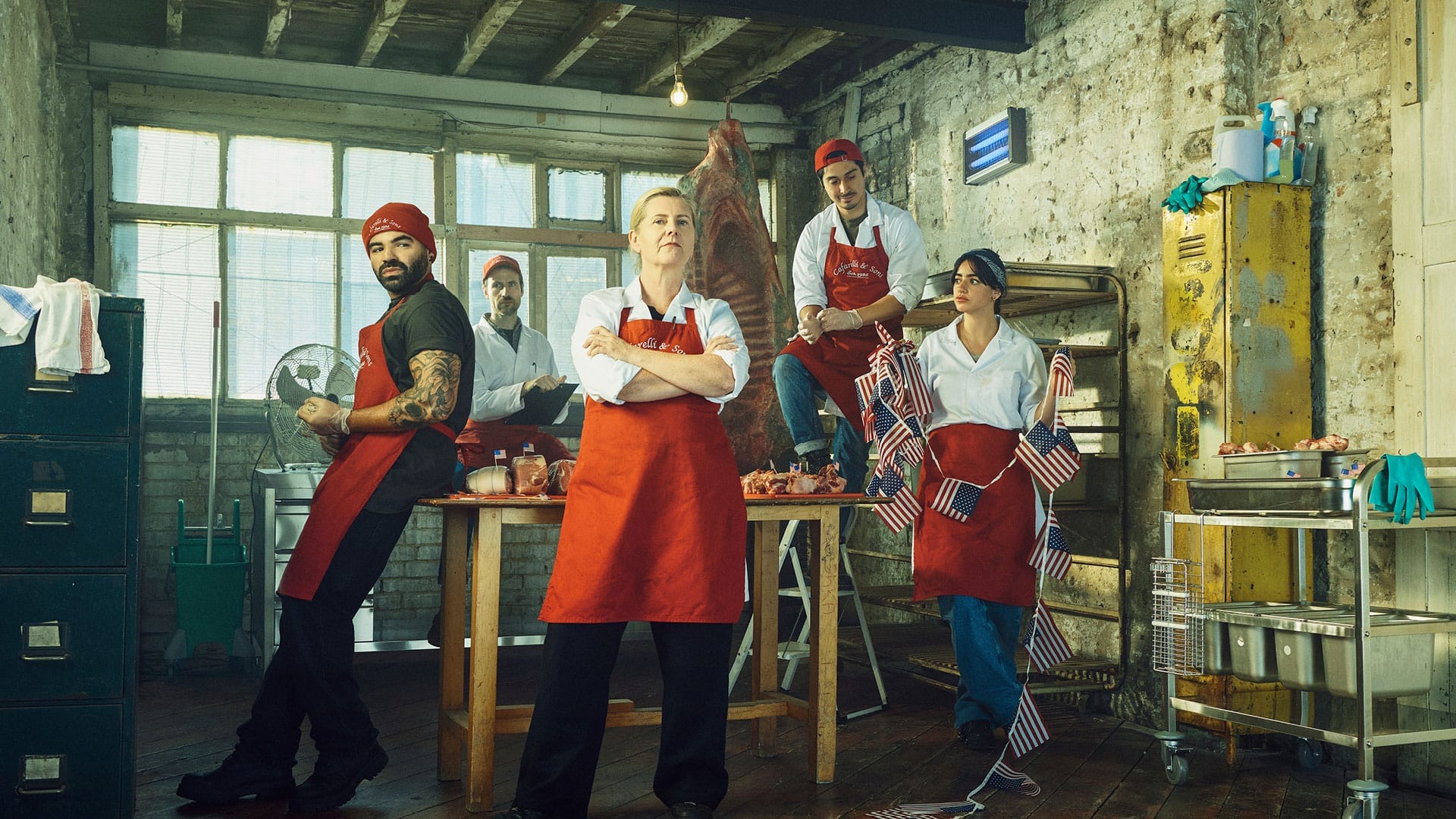 Promo poster for The Meat kings. Jackie Clune stands in the centre in front of a wooden table with meat on. She wears a red apron and white coat with her arms folded. Ash Hunter leans on the left side of the table in a red beanie, black tshirt and jeans and red apron. Mithra Malek stand to Jackie's right, holding American flag bunting. She wears a white coat and a red apron. Marcello Cruz sits on a ladder above Mithra. He wears a red backwards baseball cap, a white coat and red apron. Behind the table Eugene McCoy stands holding a clipboard. He wears a red baseball cap and white coat.