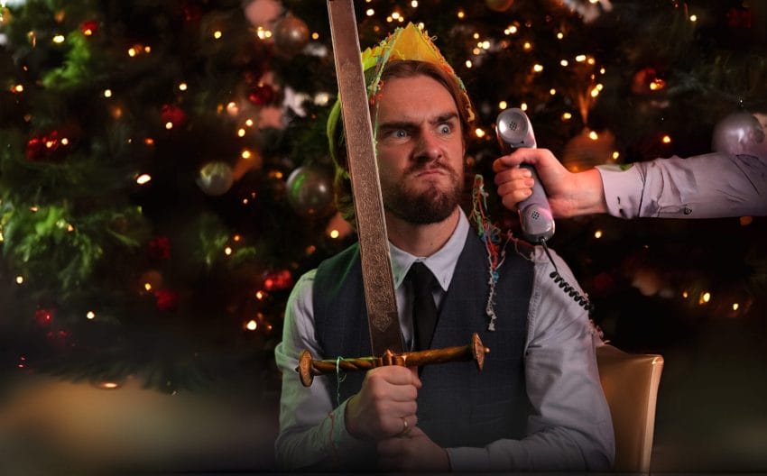 Felix Grainger sits in front of a christmas tree. He is wearing a white shirt, black tie and navy waistcoat. He holds a shord and there is a yellow paper crown on his head. He has a comically angry expression. A hand holds out a phone to him.