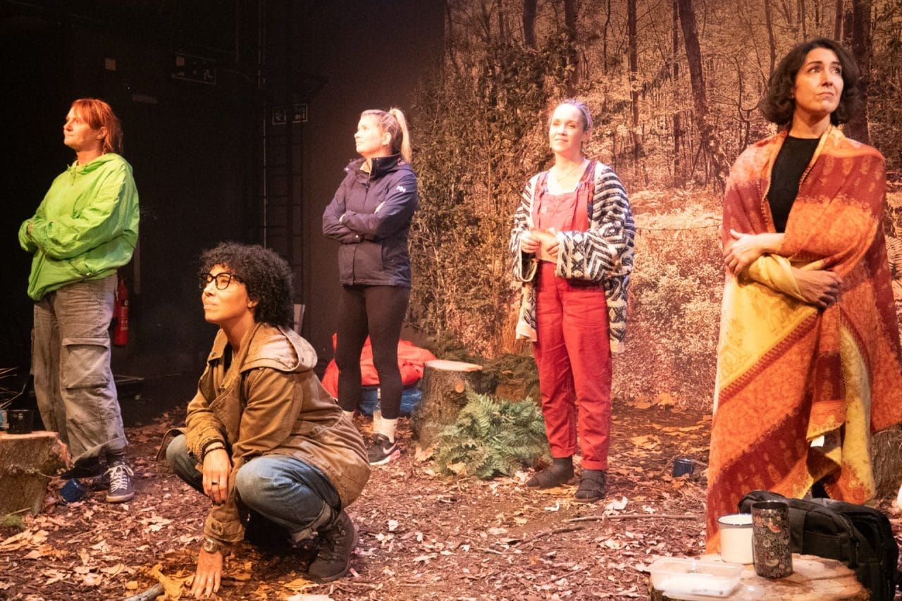 Stacy is crouching centre stage with the other women standing behind her. They are all looking in different directions and all have a peaceful expression.