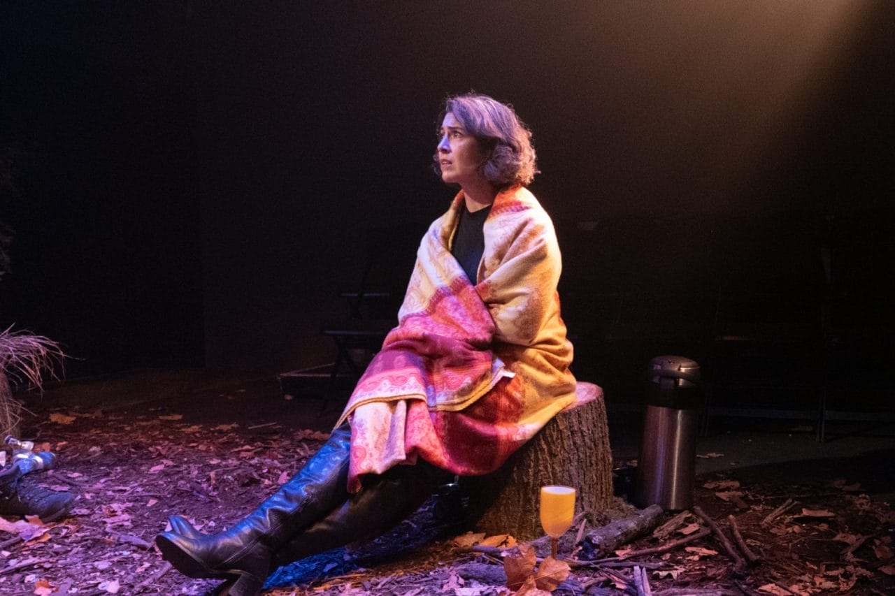 Rendah sits on a tree stump with a red and yellow blanket around her shoulders. Her legs are stretched out in front of her and she wears thigh high black boots.